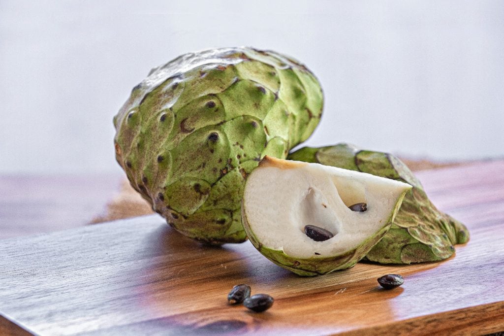 quartered cherimoya lays in front of whole cherimoya revealing white interior and dark brown seeds