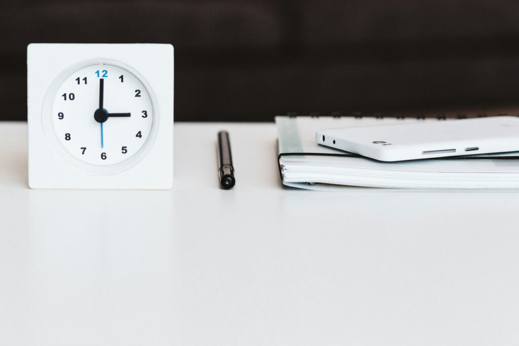 Eine Uhr auf einem Schreibtisch neben einem Stift und einem Block symbolisiert die Zeiterfassung für Kleinbetriebe.