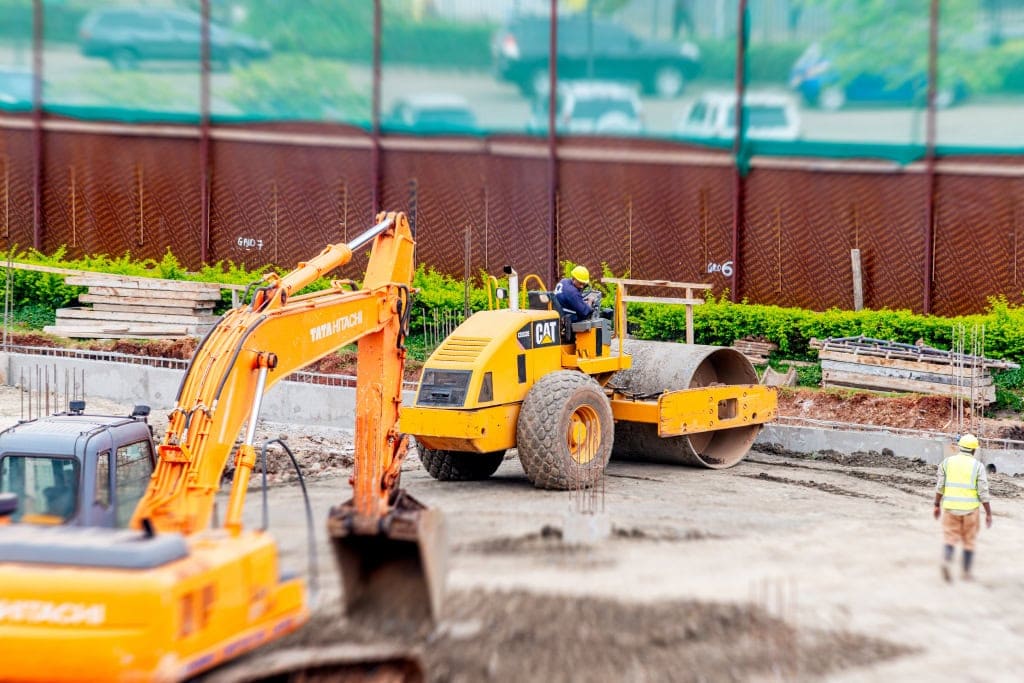 An orange excavator and yellow road roller operate at a construction site while workers stand nearby.
