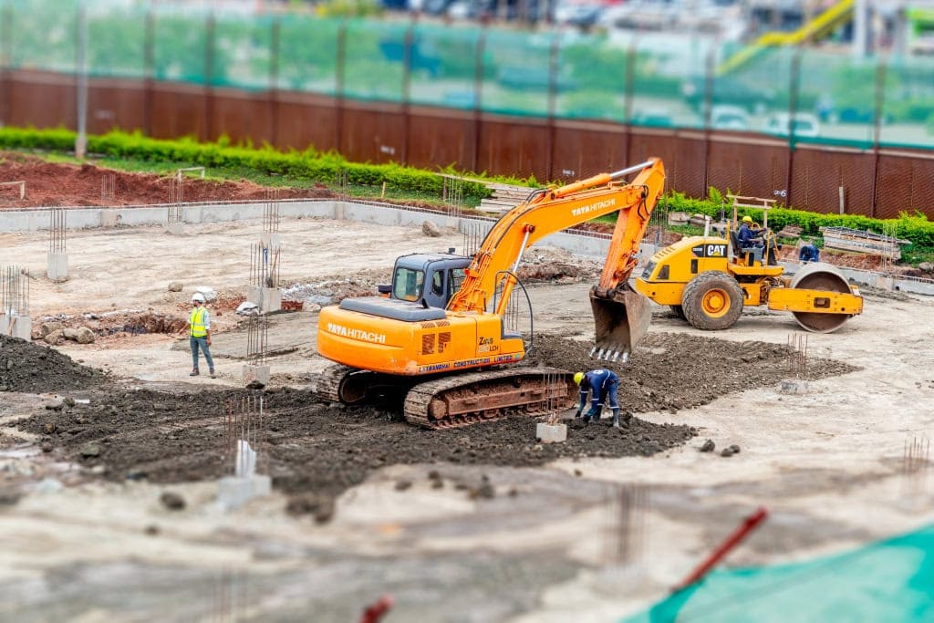 An orange excavator and yellow road roller work among workers and concrete foundation columns.