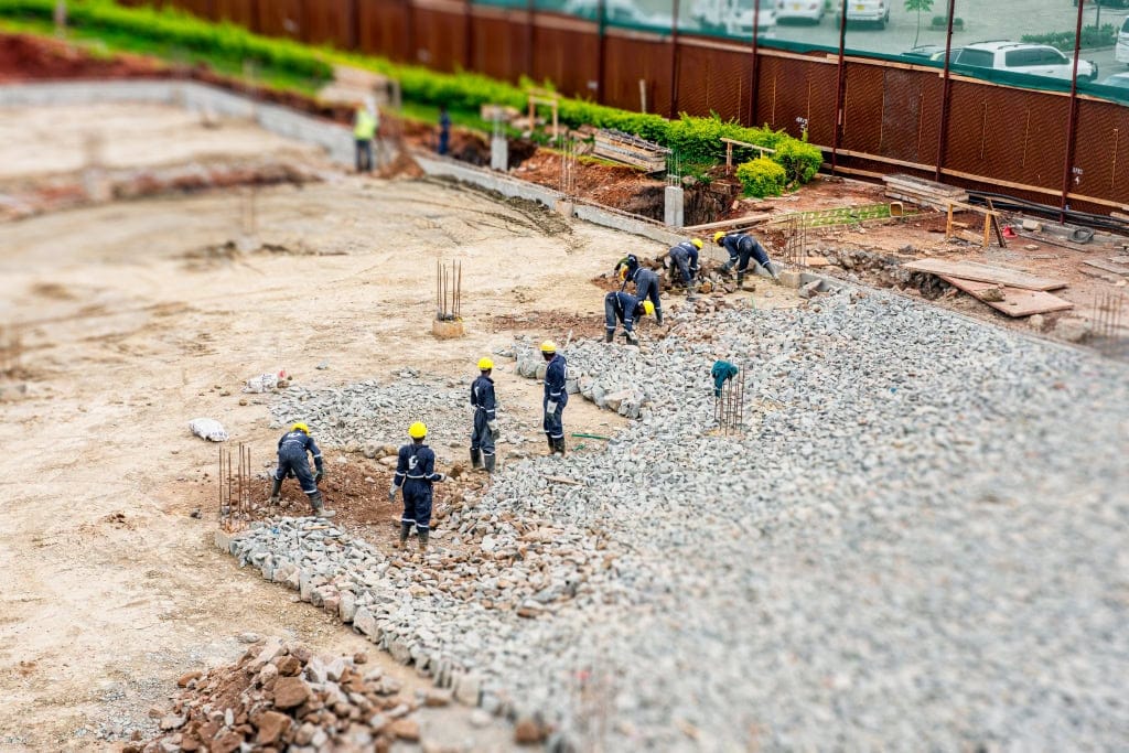 A group of helmeted workers spreads gray aggregate across a construction foundation area.