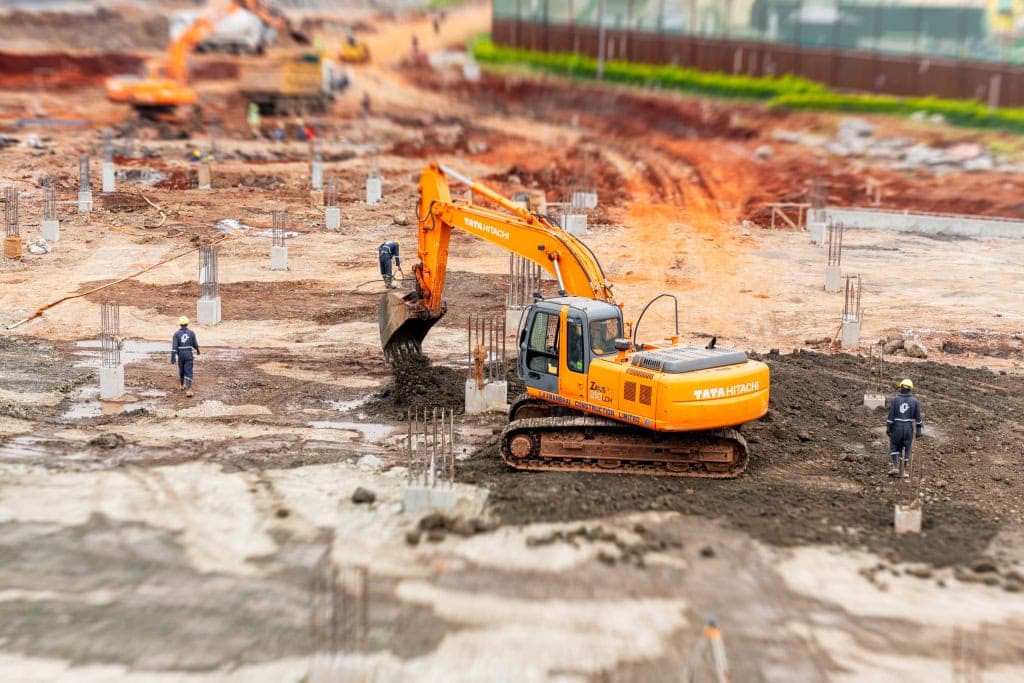 An orange excavator lifts soil from a trench while workers and reinforced foundation columns surround it.