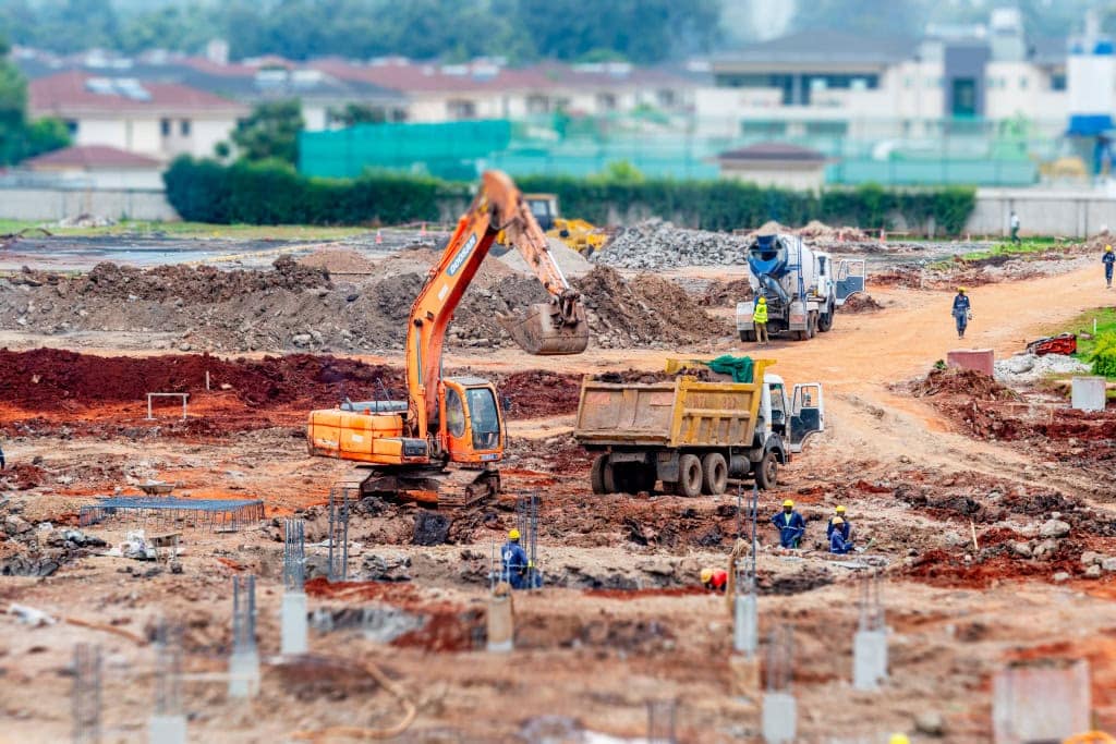 An excavator, dump truck, and concrete mixer operate across a large construction site with workers nearby.