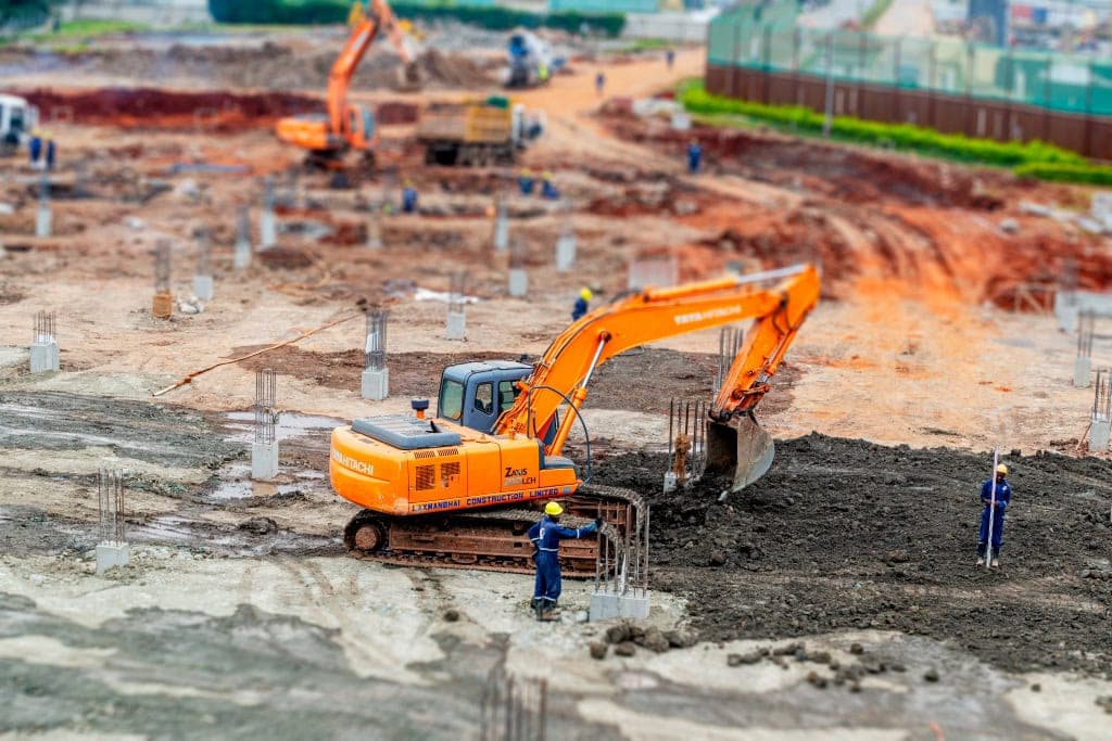 Orange excavator operates among workers and concrete foundations at a construction site