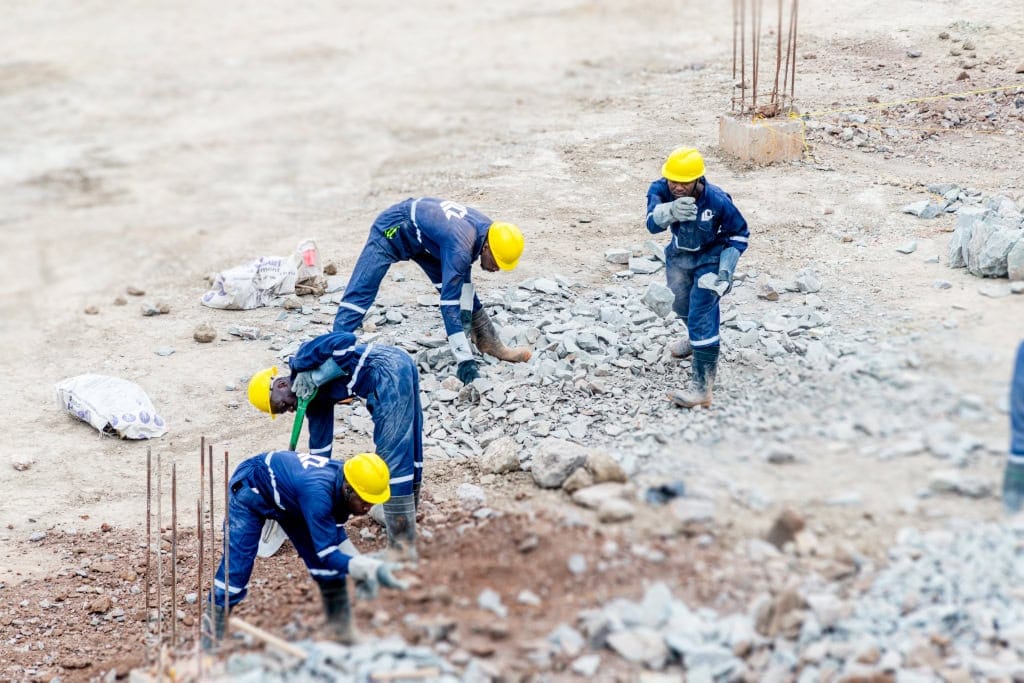 Construction workers handle rocks and soil around exposed foundation reinforcement
