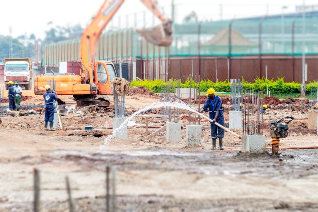 A worker sprays water across foundation columns while excavators operate behind him