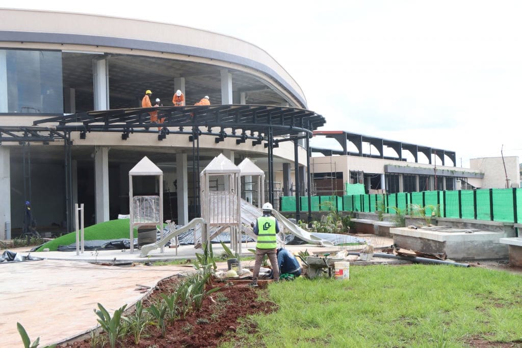 Workers install equipment beside a playground and landscaped lawn outside a mall building.