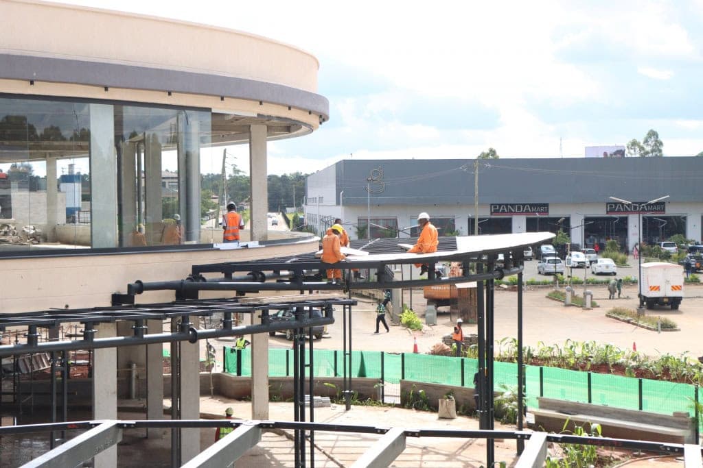 Workers in orange clothing work on a canopy beside a rounded mall building.