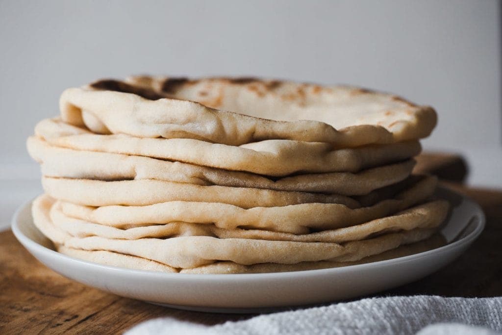 El Bazlama es un increíble y delicioso pan plano turco, suave y acolchado! Es extra tierno y hueco, un mix perfecto entre el pan Naan y el pan Pita. Sírvelo con tus salsas favoritas, carne asada, hummus o disfrútalo solo tal cual. Foto frontal de bazlamas en una pila.