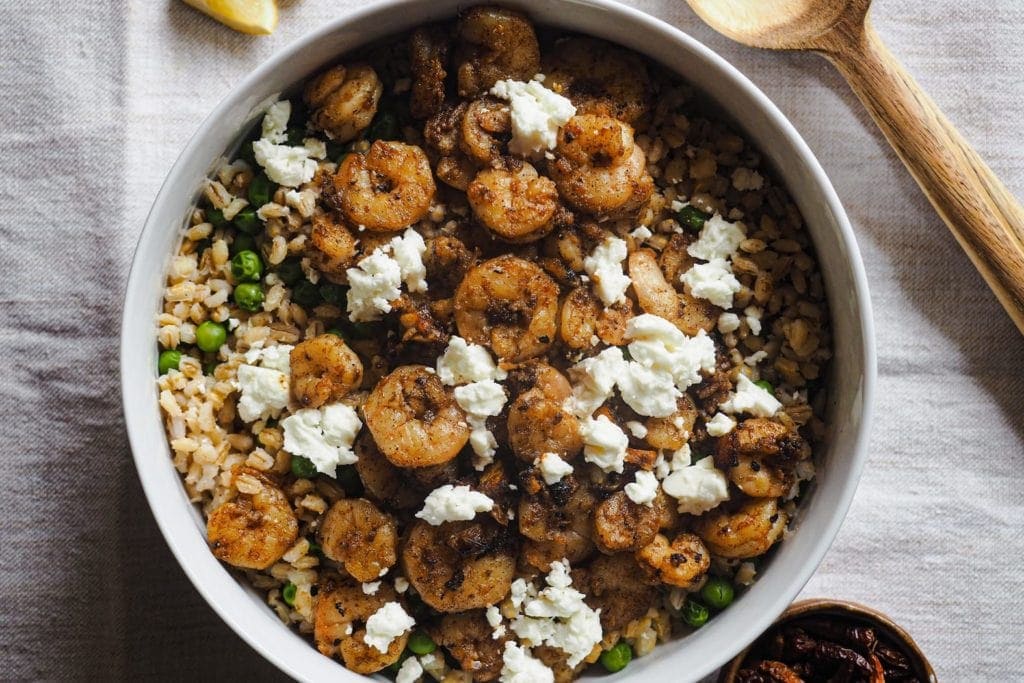 Ensalada Tibia de Gambas y Cebada: súper deliciosa, nutritiva y fácil de preparar. Tiene toques de limón, ajo y pimentón, con guisantes y feta, que se deshace al mezclarlo con las gambas calientes para redondear el plato. En esta foto cenital se ve la ensalada servida en un bol con gajos de limón arriba a la izquierda, cubiertos de madera para servir la ensalada arriba a la derecha y un pequeño bol con guindillas abajo a la derecha.