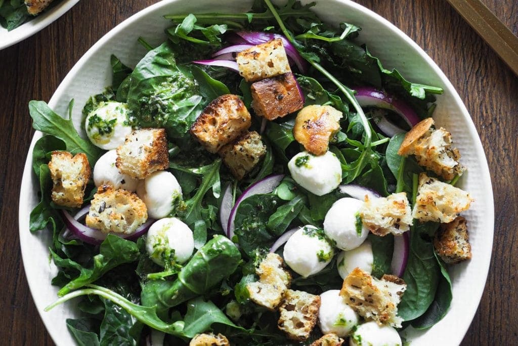 Esta ensalada de espinacas con vinagreta de alcaparras combina todos los ingredientes clave de una buena ensalada: verde del bueno con espinacas y rúcula, crujiente con picatostes, color con el contraste blanco de la mozzarella y el morado de la cebolla, todo ligado con una deliciosa vinagreta de alcaparras. Si tienes picatostes a mano, en 5 minutos la tienes lista! Foto desde arriba en horizontal de un plato con la ensalada.