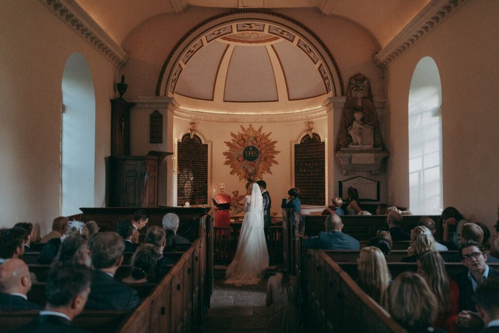 A bride and groom stand at the altar in a small, warmly lit church, as a Creative Wedding Photographer captures the moment beneath a domed ceiling and a large golden religious symbol, surrounded by friends and family.