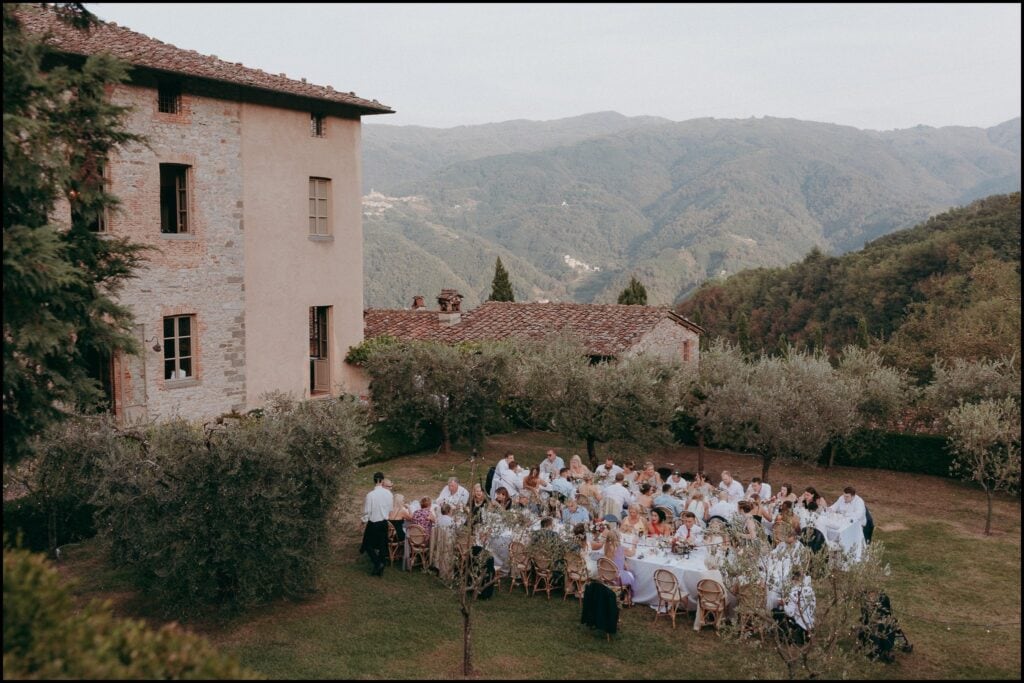 A group of people sits at tables set for an outdoor meal in a garden next to a stone building, surrounded by trees and hills under a slightly overcast sky, perfectly captured by a creative wedding photographer.