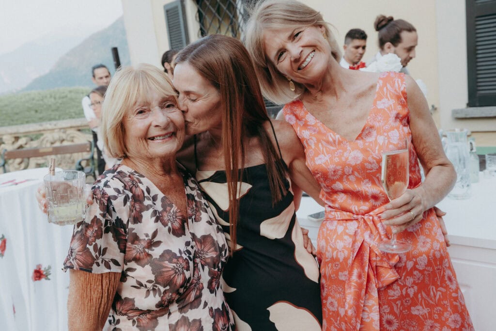 Three smiling women pose together at an outdoor event. The woman in the middle kisses the woman on the left on the cheek. They are dressed in elegant, floral-patterned dresses and hold drinks, with guests and greenery in the background.