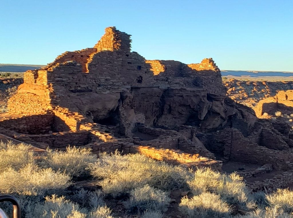 Three-story ruins made of shaped rock reflect the setting sun in a wide open, dry environment