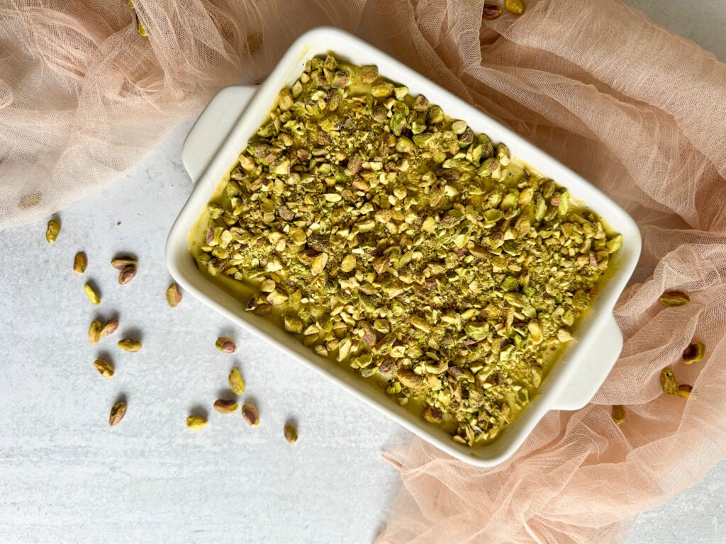 Pistachio tiramisu in a white baking tray over a table with a pink cloth and some pistachios.