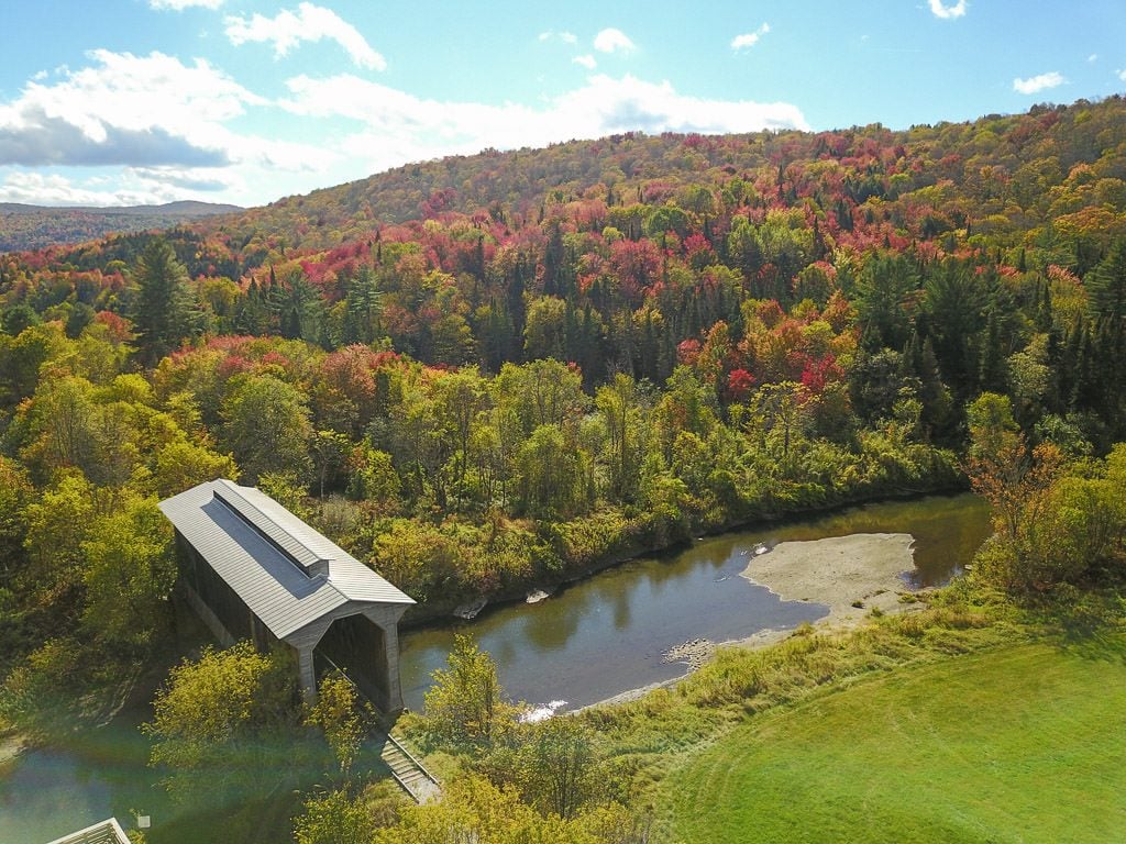 Drone shot of a covered bridge going over a river and surrounded by bright and colorful trees in vermont during fall