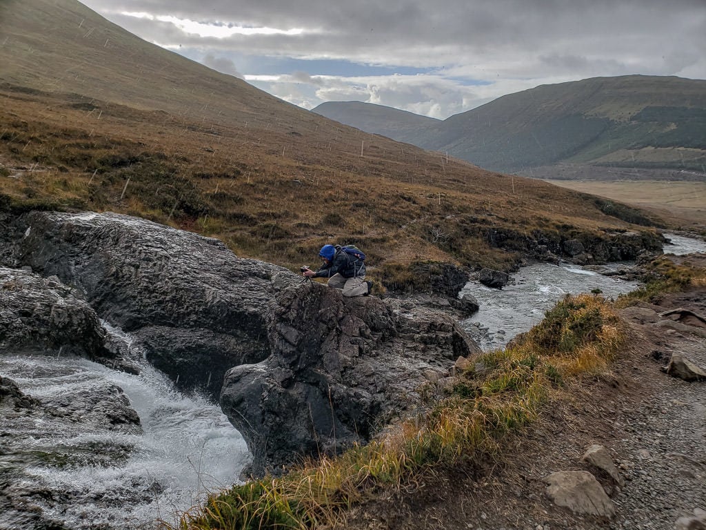 Buddy kneeling on a rock overlooking a waterfall as he takes a photo in the heavy rain during our visit to Fairy Pools in Isle of Skye