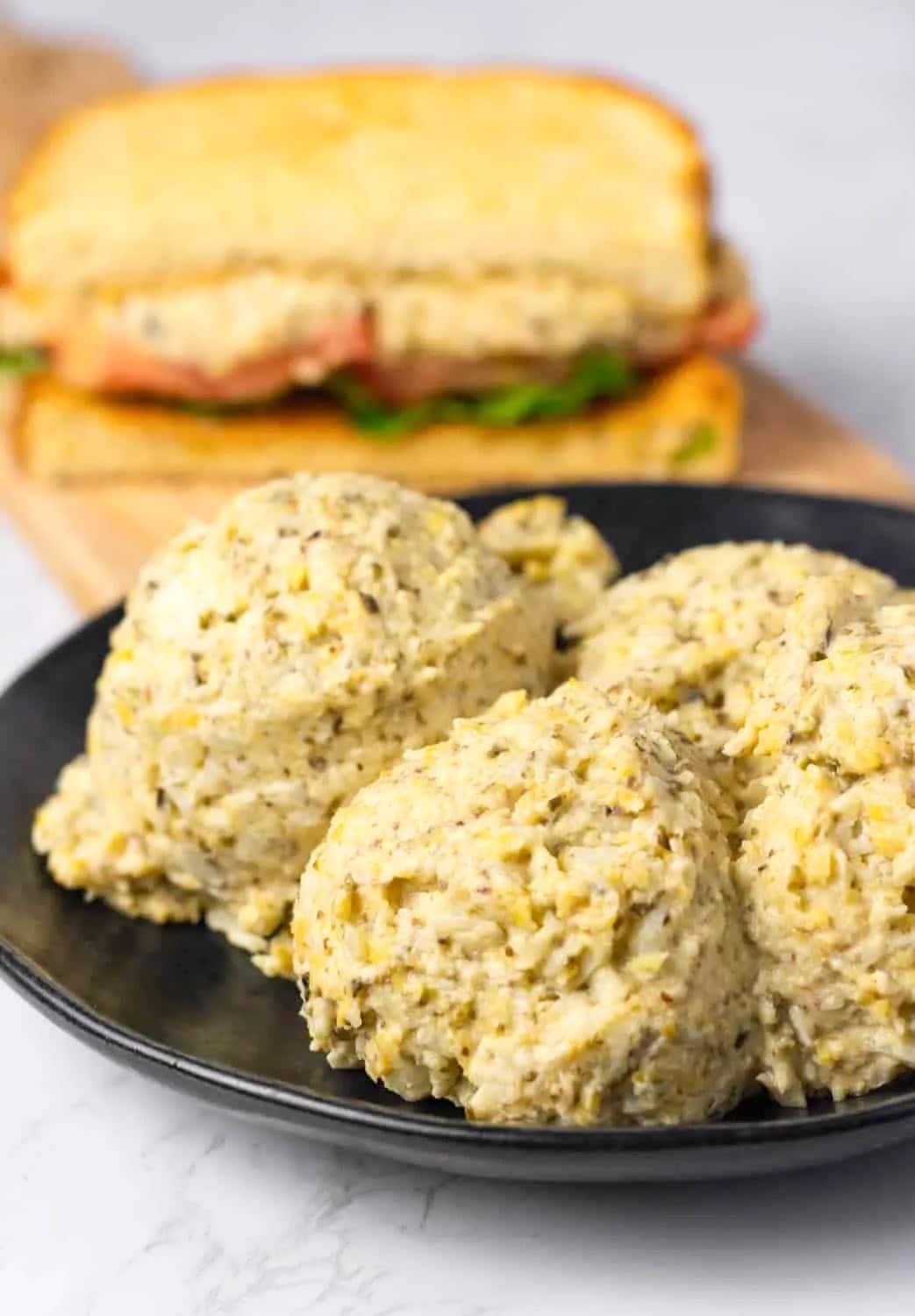 3 scoops of tuna salad on a black plate with a tuna sandwich in the back on a wooden cutting board on a white background