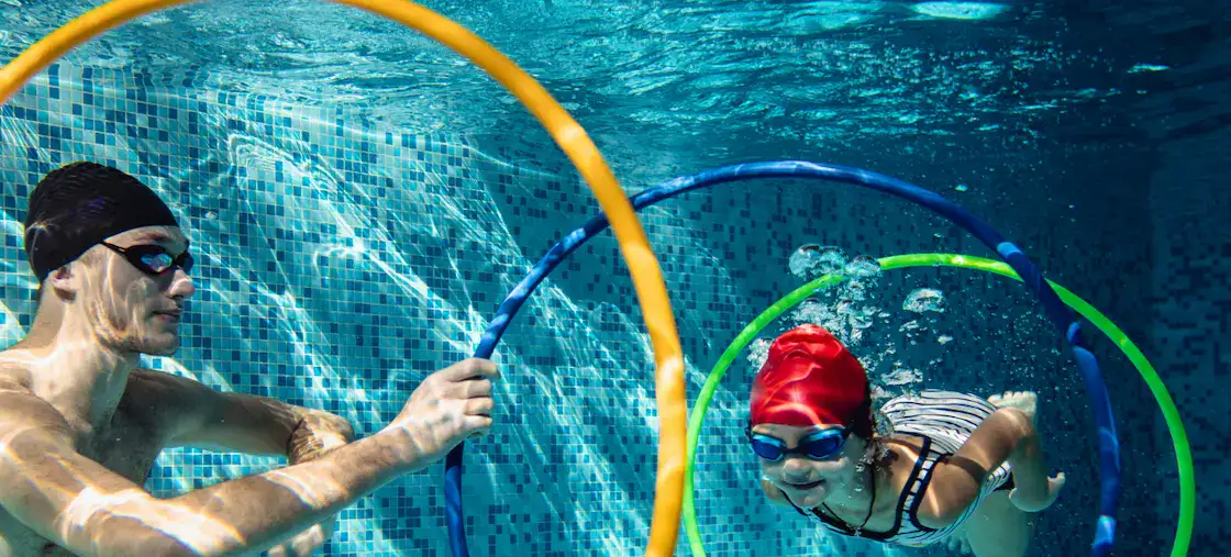 school-aged child is confidently swimming under the water in a swimming pool through the hoops with swim teacher holding a hoop in a private swim lesson at Blue Wave swim school in Wimbledon