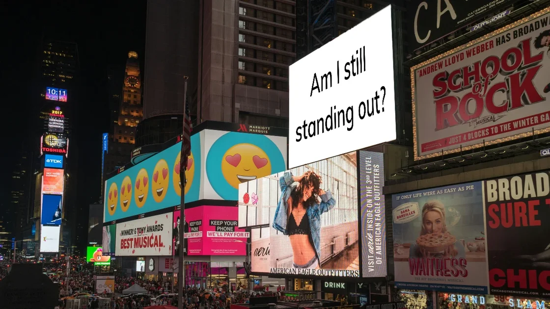View of busy inner-city intersection with lots of billboards. One billboard reads "Am I still standing out?"