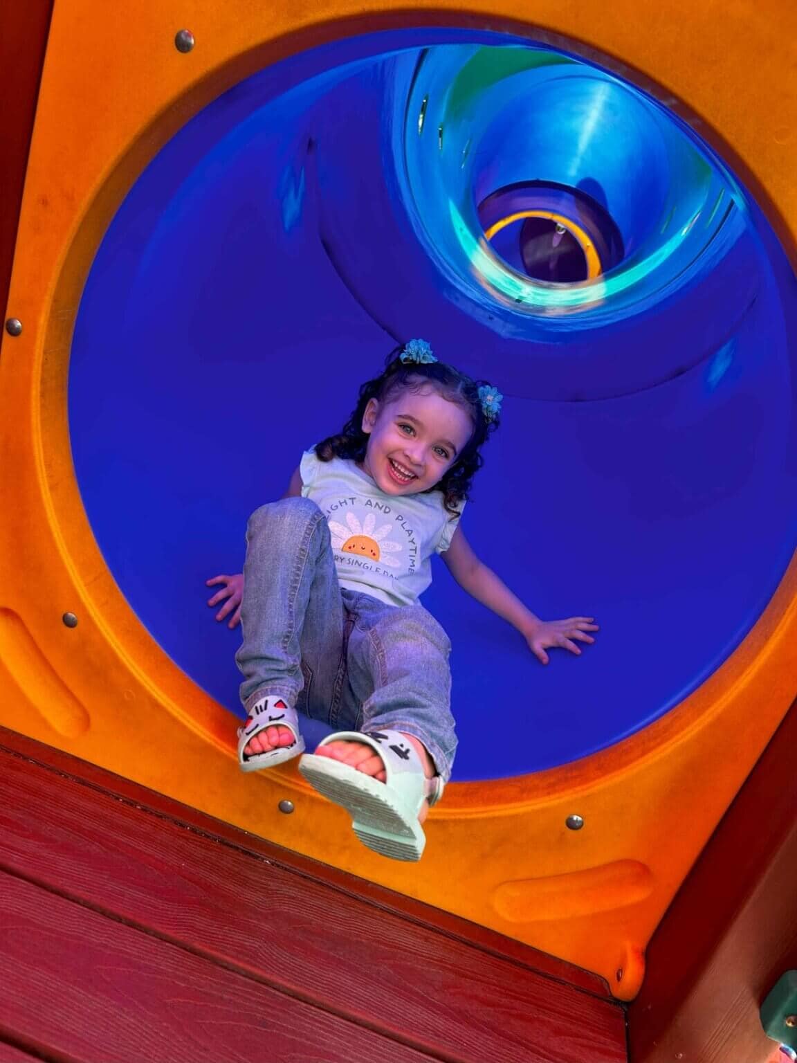 Menina sorridente brinca em escorregador de parque infantil rosa e azul, demonstrando diversão e alegria em um dia ensolarado ao ar livre - kreke Pernambuco
