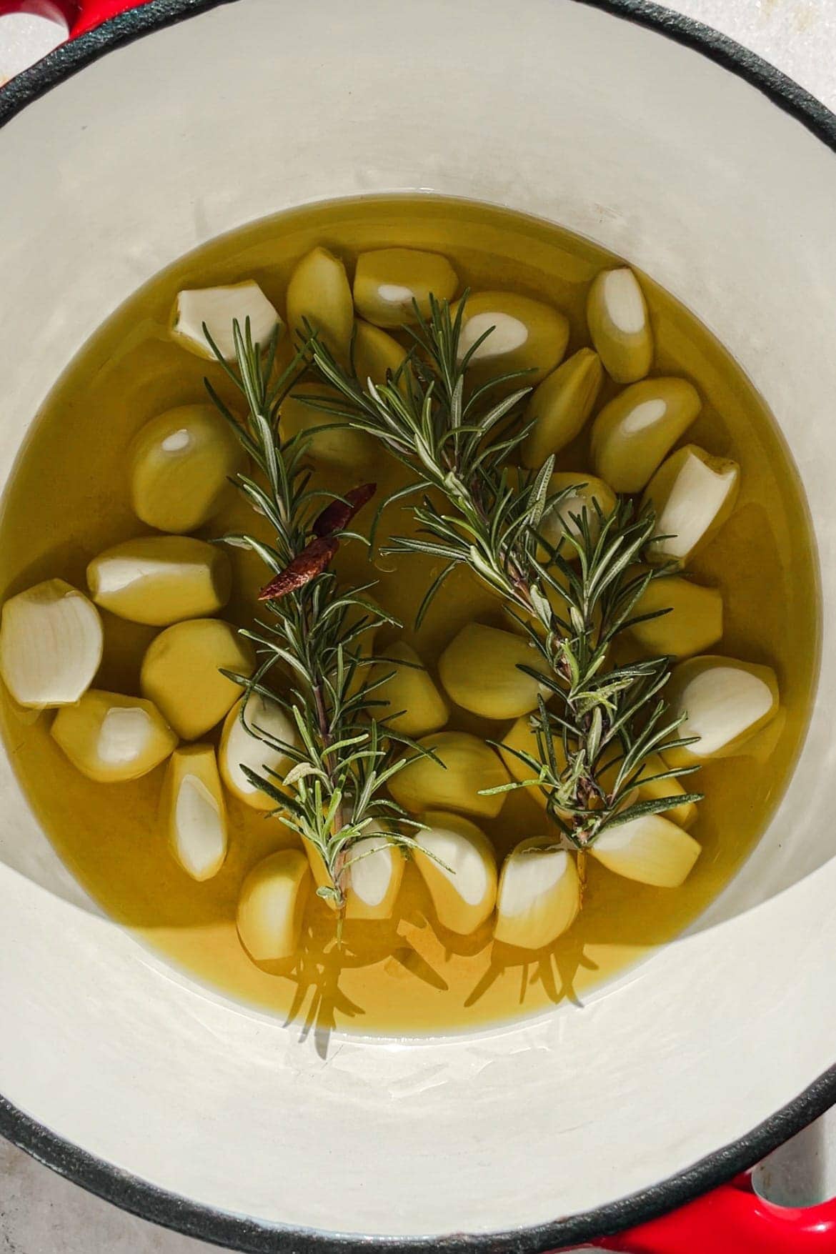 This Garlic Confit recipe is going to become your new pantry staple because it's so easy and confited garlic is so spectacular and versatile. You just need a few heads of garlic, your favourite spices and herbs, lots of olive oil and 30 minutes. The result is a scrumptious sort of sweet spreadable garlic that you can use in a thousand ways. As a bonus, you get garlic-infused oil! In this picture you can see the raw garlic in a cocotte with chillies and rosemary covered in olive oil.| Paula's Apron Recipe