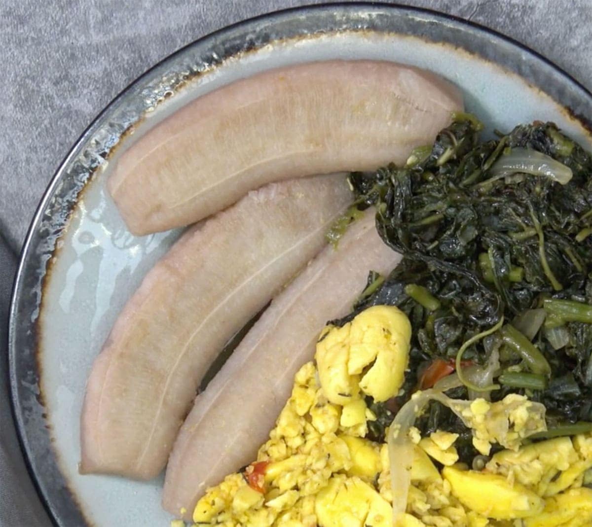 boiled green banana on a blue plate with ackee and callaloo on a grey background