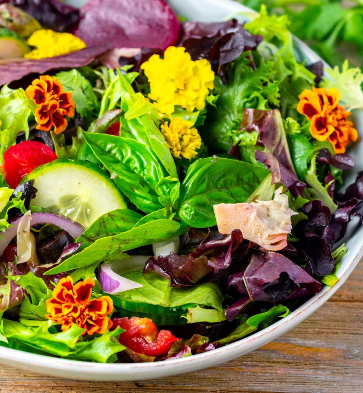 close up of spring salad mix in a blue bowl, basil, edible flowers on a wooden board