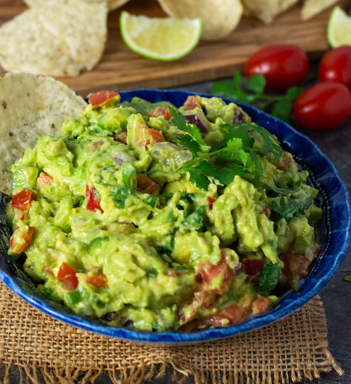 Loaded guacamole recipe with cilantro leaves, tortilla chips on a grey background with cutting board and extra ingredients in the background