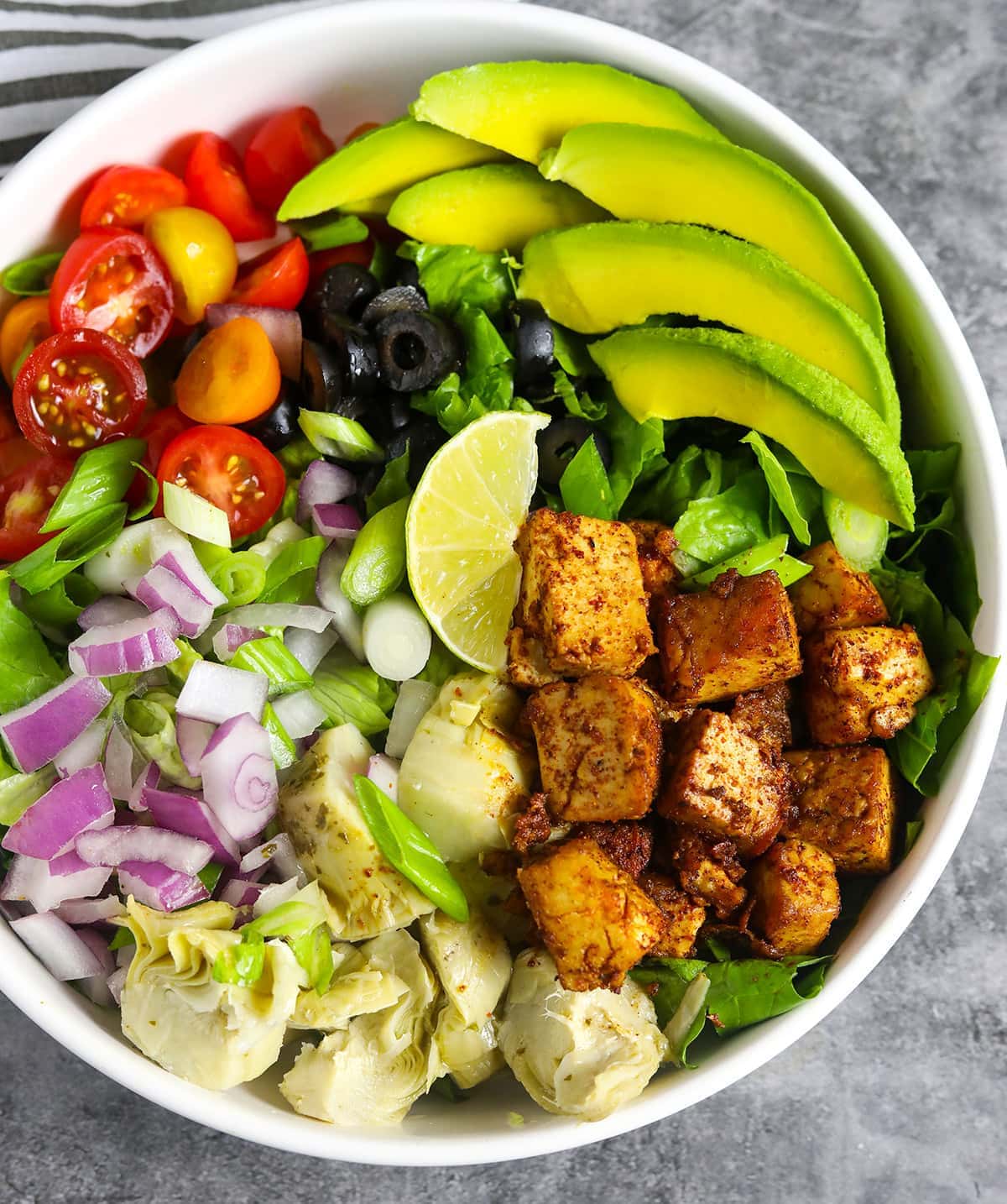 Overlay Cajun tofu salad in a white bowl with slices of avocado, tomato, red onion, marinated tofu, artichoke hearts, green onion, lettuce, olives in a white bowl on a grey background