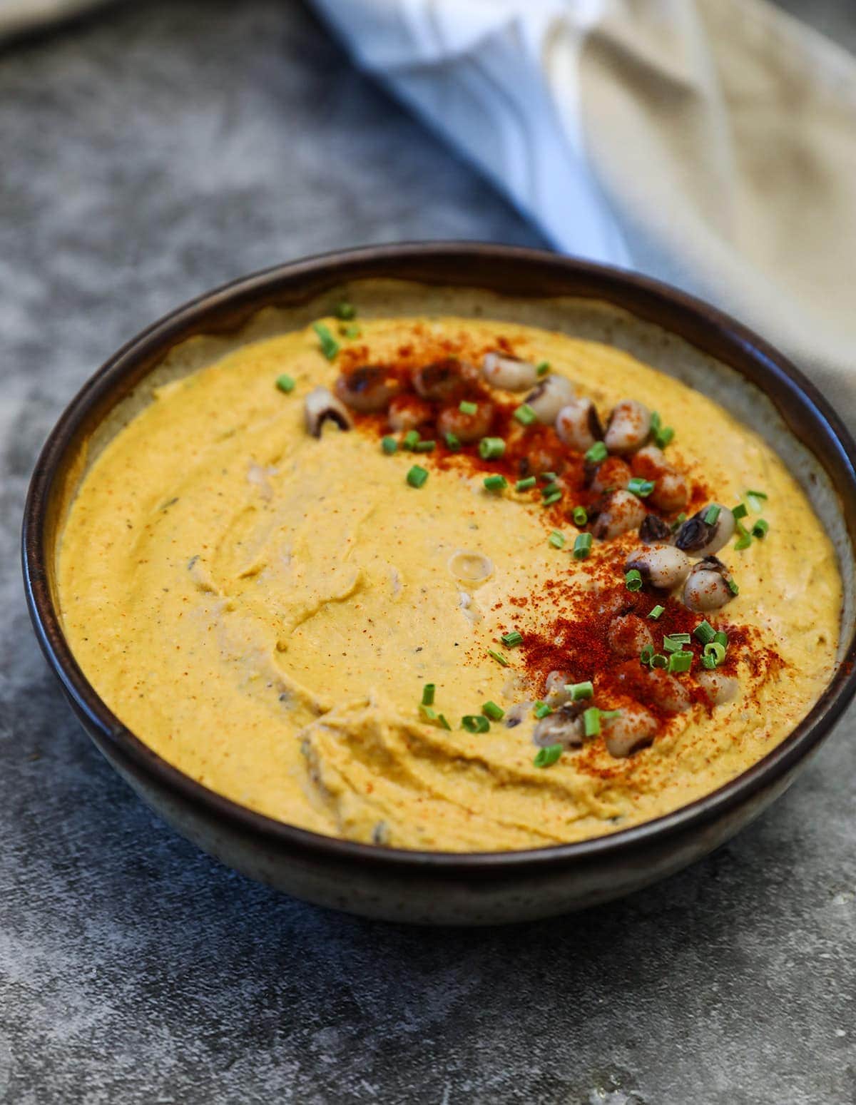 black eyed peas hummus in a brown bowl on a grey background garnished with black-eyed peas and paprika