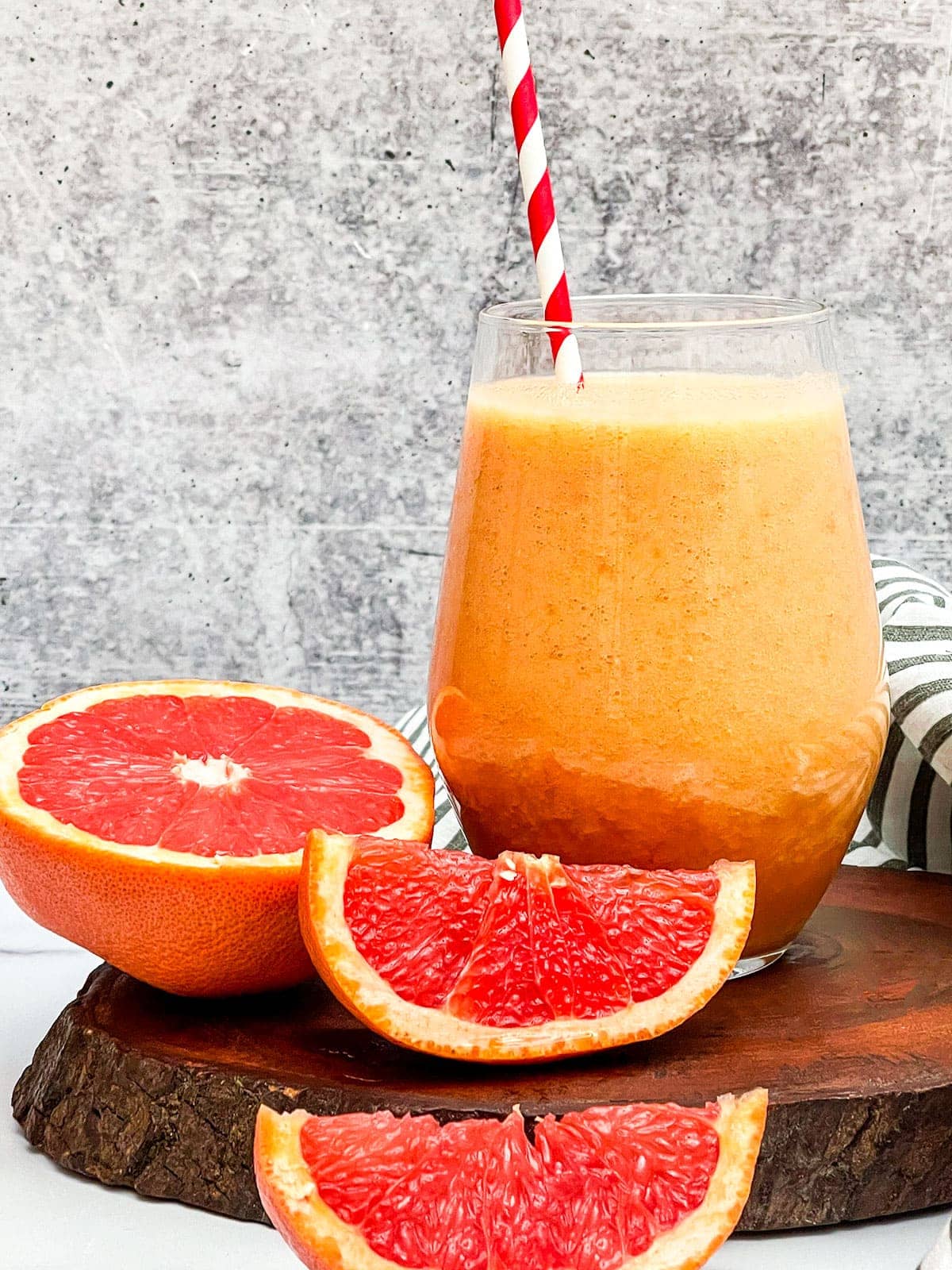 Grapefruit smoothie in a glass with a striped orange and white straw, cut pieces of grapefruits on a wooden background