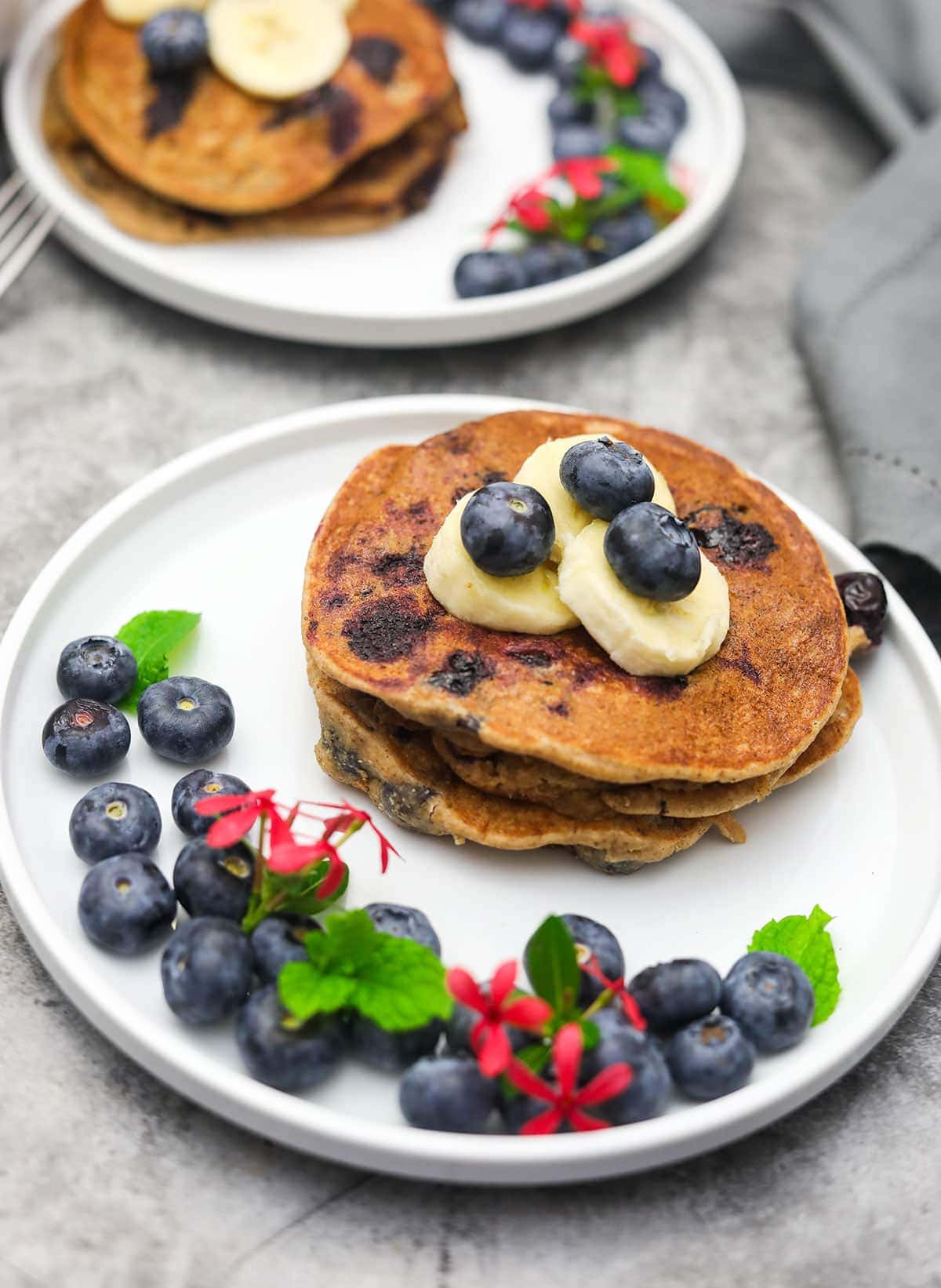 blueberry banana oatmeal pancakes stacked on two white plates decorated with blueberries, banana slices and pink flower on a concrete background