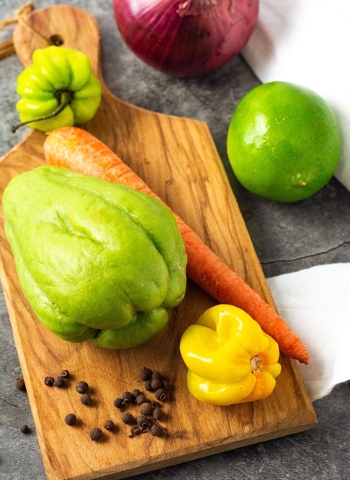 Carrot, chocho, onion, scotch bonnet pepper, pimento and lime on a cutting board on a grey background