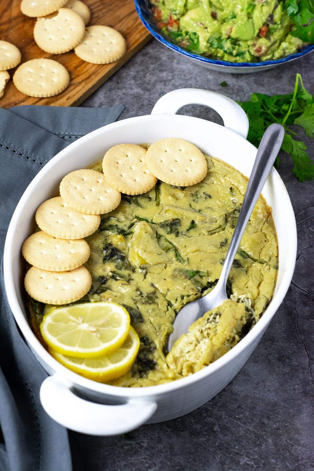 Vegan spinach dish overlay, topped with gluten-free vegan crackers and serving spoon in a white casserole dish on a grey background