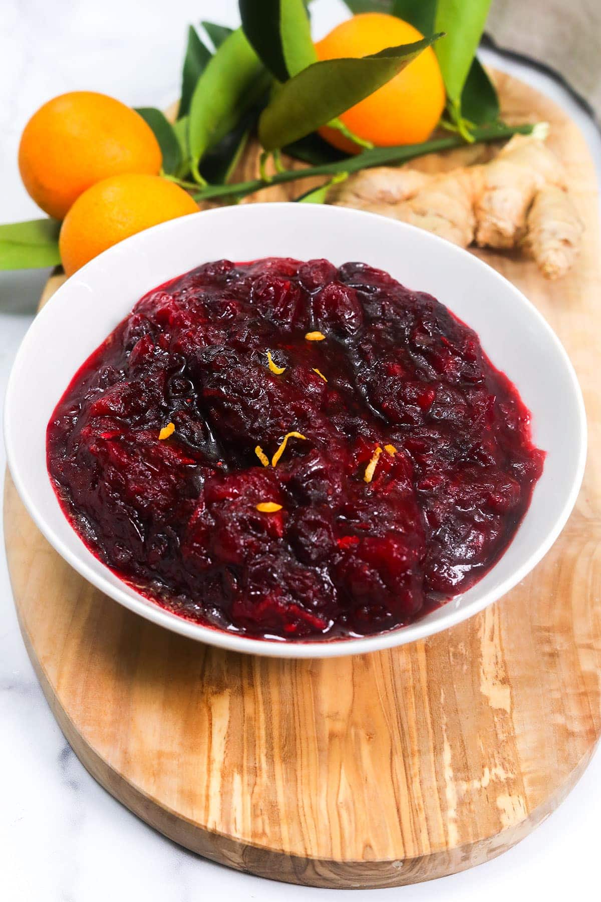 Vegan cranberry sauce in a white bowl on a cutting board with oranges and ginger in the background