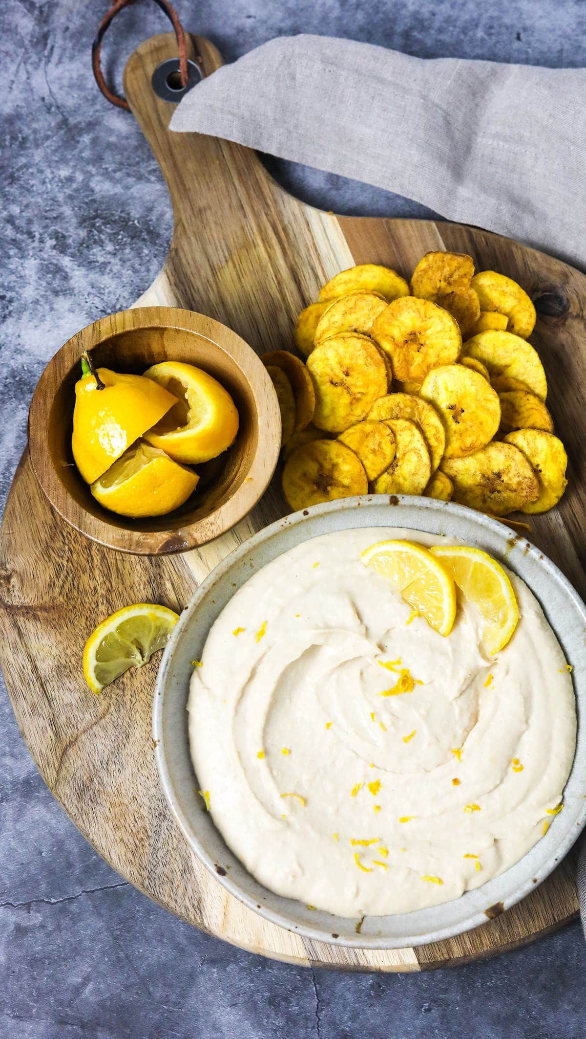 overlay of hummus in a beige speckled bowl on a wooden background with banana chips and lemon in a wooden bowl