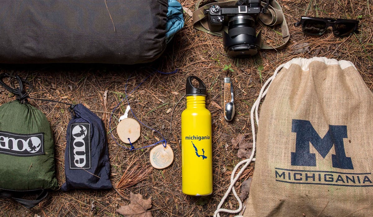 Camp Michigania bag and water bottle on the ground with sunglasses and a camera