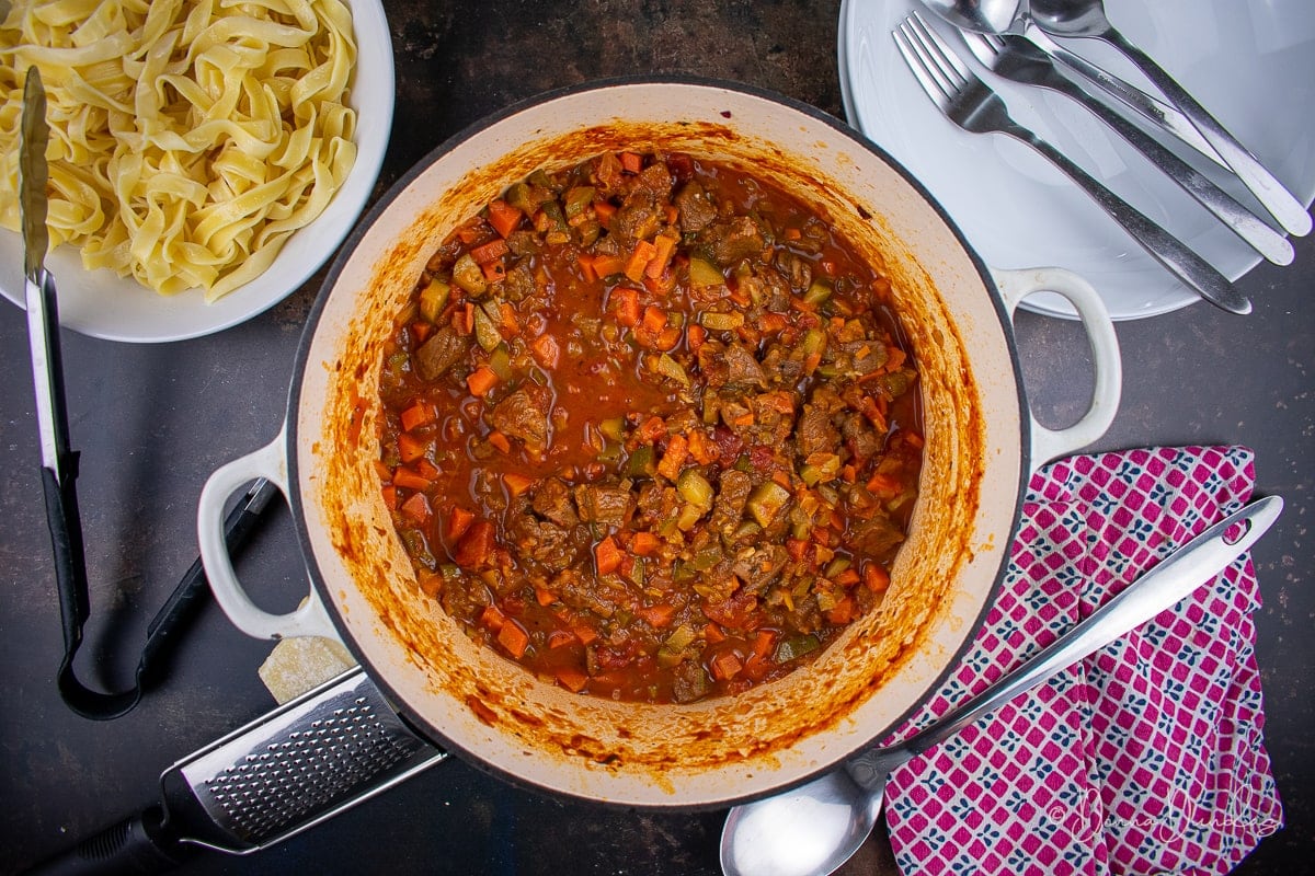 cooked ragu in a large cast iron dish. White bowl filled with pasta to the side. Empty bowls with spoons and forks to other side. Serving spoon, red and blue patterned towel at the bottom.