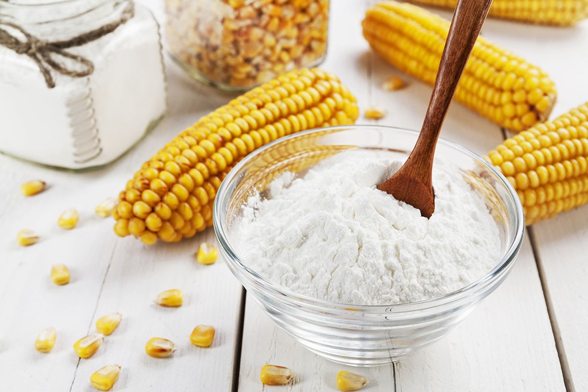 cornstarch in a glass bowl with a wooden spoon on a white background with dried corn ears and kernels. A great Arrowroot Flour Substitute