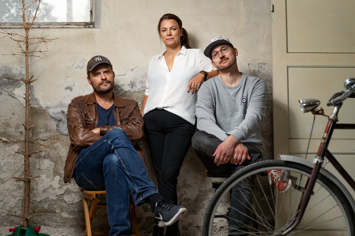 Red Carpet Media Gruppbild i rörig miljö. Två män sitter ner på stolar, och en kvinna står mellan dom. I bakgrunden syns ett fönster, en barrlös julgran, och i förgrunden en cykel.