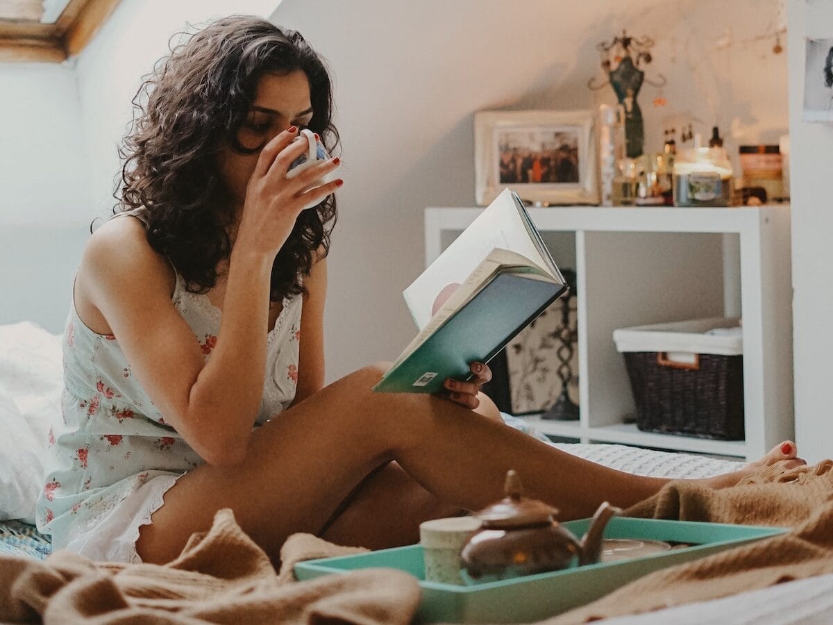 woman sitting on bed reading a book and drinking coffee