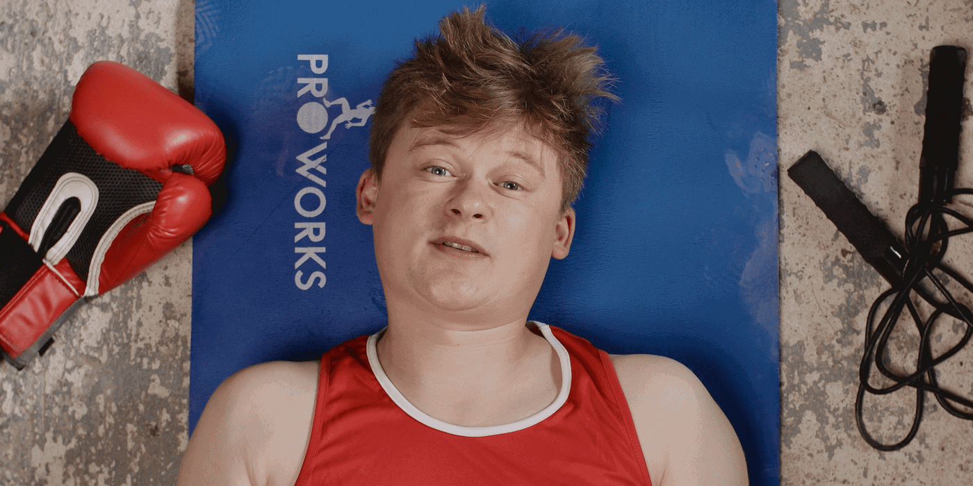 Photo of video essayist Tom Nicholas, head and shoulders shot of him lying on the floor looking at the camera, a boxing glove to one side of his head and a jump rope to the other.