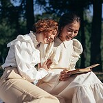 smiling women in old fashioned clothing reading book on picnic blanket