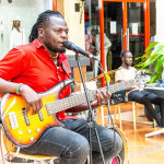 Musician in a red shirt singing into a microphone while playing bass guitar