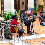 Band performing with drums, guitars, keyboards, and saxophone in a shopping mall