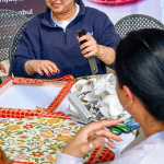 A facilitator demonstrates a crafting tool while a participant follows along at the Frame and Fabric Workshop.