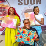 Three participants display colourful fabric artwork at the Frame & Fabric Workshop, with scissors and materials visible on the table.