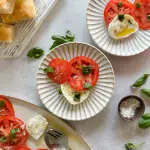 Caprese Salad on scalloped whit plate with breads and basil leaves on the table!